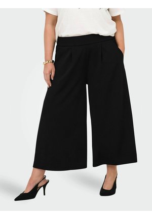 Person med vide svarte kortbukser, hvit topp, svarte slingback hæler og et gullarmbånd på venstre håndledd.