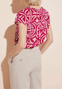 Růžová halenka s krátkými rukávy a geometrickým bílým vzorem. Má kulatý výstřih a záložku na zádech. Kombinováno se světle šedými kalhotami.