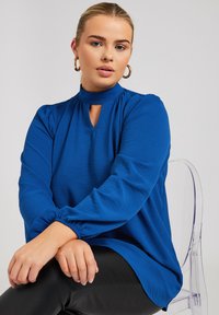 Blusa azzurra a maniche lunghe con collo alto, dettaglio a chiave e maniche arricciate, realizzata in tessuto testurizzato. Abbinata a pantaloni neri.