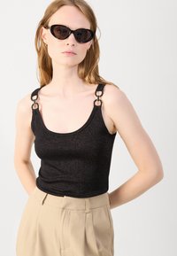 Black sleeveless top with a scooped neckline, sparkling texture, and metal accent details on the shoulder straps; paired with beige trousers.