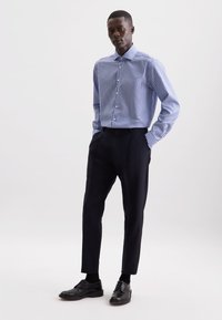 Uomo con una maglietta a righe blu e bianche a maniche lunghe infilata in pantaloni scuri slim-fit, con scarpe nere eleganti, in piedi con le mani in tasca.