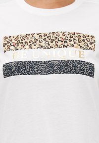 T-shirt blanc présentant trois bandes horizontales : la bande du haut en imprimé léopard avec des accents roses, la bande du milieu avec "SOIS UNIQUE" en doré, et la bande du bas en imprimé léopard noir.