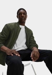 Giacca bomber verde oliva con polsini a coste, indossata sopra una maglietta bianca e abbinata a pantaloni scuri. Design semplice, texture liscia, stile pratico.
