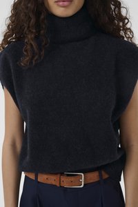 Kobieta ubrana w czarny bezrękawnik z golfem, włożony w granatowe spodnie z brązowym zamszowym paskiem, z kręconymi włosami opadającymi na ramiona.