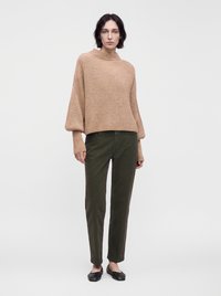 Femme portant un pull côtelé beige, un pantalon vert foncé et des chaussures plates noires, debout devant un fond clair uni.