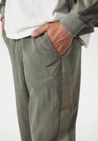 Man in olijfgroene Guess-broek met zijstreep en logopatch, hand in de zak, gecombineerd met een wit shirt en bijpassende jasje mouwen.