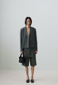 Dark green tailored suit with a blazer and shorts; smooth fabric, single-button closure, paired with a black faux fur bag and flat shoes.