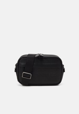 Schwarze Leder-Crossbody-Tasche mit verstellbarem Riemen, Reißverschluss und geprägtem Markennamen auf der Vorderseite vor weißem Hintergrund.