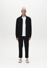 Chaqueta negra con cremallera sobre una camiseta blanca, combinada con pantalones negros y zapatillas gris claro, rematada con un gorro de punto beige claro.