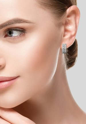 Boucles d'oreilles en argent en forme de boucle, composées de trois bandes empilées, avec une surface texturée qui reflète la lumière, portées sur l'oreille d'un modèle.