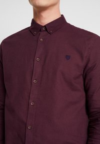 Man in een dichtgeknoopte, langekleurige, donkerburgundy blouse met een button-down kraag en een klein geborduurd logo op de borst.