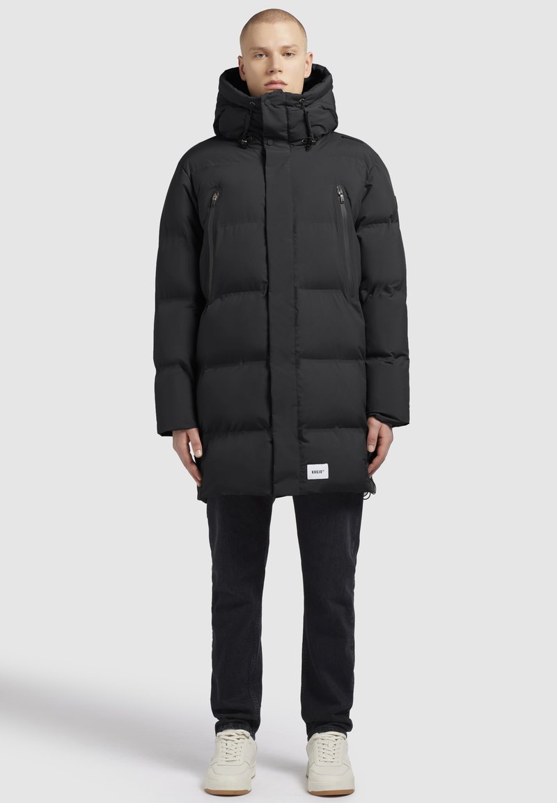 Svart puffjacka med hög krage, framfickor med dragkedja och en logotyp-plåster. Mjuk textur och vadderad design för isolering.