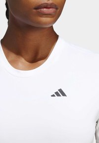 Vit idrottsskorta med rundad halsringning och subtil textur, prydd med en liten svart tre-stripes-logotyp på vänster bröst.