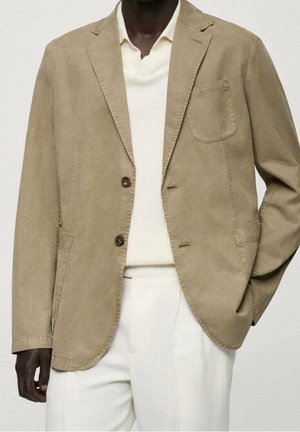 Mies, joka pukeutunut beigeen bleiseriin valkoisen neulepaidan ja valkoisten housujen kanssa, toinen käsi taskussa, yksiväristä taustaa vasten.