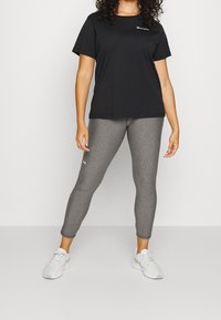 Kobieta ubrana w czarny T-shirt Champion, szare legginsy Under Armour oraz białe sneakersy, stojąca na jednolitym tle.