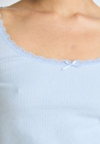 Svetlo modra majica na bretelice z izrezom v obliki valov, z majhno pentljo na sredini in teksturiranim rebrastim vzorcem.