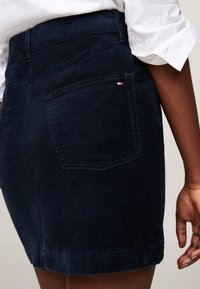 Primer plano de una persona que lleva una falda de pana azul marino con bolsillos traseros y un pequeño logo rojo, blanco y azul en el bolsillo derecho.