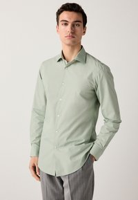 Lysegrøn langærmet skjorte med knaplukning på kraven, glat tekstur, knapper foran og buet kant, kombineret med grå stribede bukser.