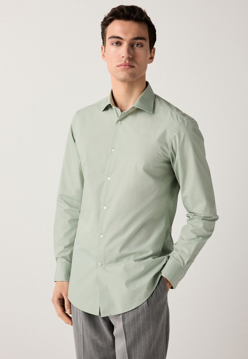 Lysegrøn langærmet skjorte med knaplukning på kraven, glat tekstur, knapper foran og buet kant, kombineret med grå stribede bukser.