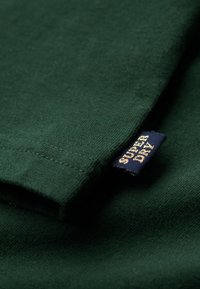 Grøn bomulds t-shirt med glat tekstur; har en foldet ærme og et blåt mærke med guldtekst, der viser brandnavnet.