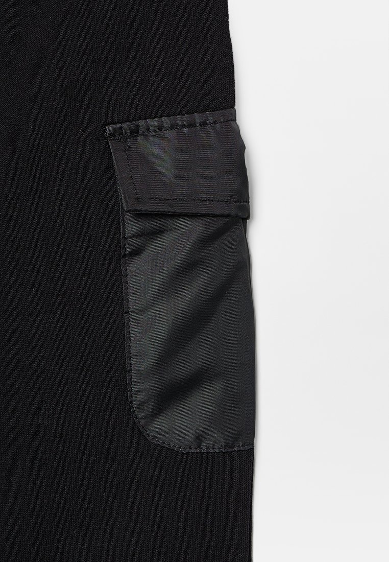 Tissu noir avec un détail de poche lisse et brillant en tissu contrasté. La poche présente un rabat replié et est soigneusement piquée.