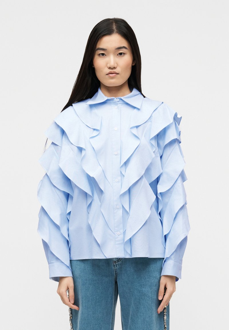 Νέα γυναίκα με μακριά σκούρα μαλλι ά, που φοράει ένα ανοιχτό μπλε μπλουζάκι με βολάν και μπλε τζιν, σε απλό λευκό φόντο.