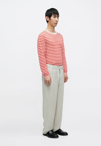 Chemise à manches longues rayée rouge et blanche associée à un pantalon beige clair et des chaussures noires. Le mannequin se tient devant un fond blanc.