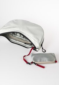 Graue wasserdichte Tasche mit abgerundetem oberen Teil, schwarzem Reißverschluss und rotem Riemen. Beinhaltet ein kleineres graues Täschchen mit rotem Riemen und Reißverschluss.