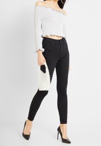 Top crop gris de canalé con hombros al descubierto y bordes acampanados, combinado con jeans skinny negros y tacones de punta negra. Bolso de cuentas blancas en la mano.