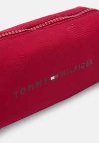 Estojo de lápis vermelho em nylon com uma superfície texturizada, apresentando um logo branco e azul. Inclui um fecho de zíper na borda superior.