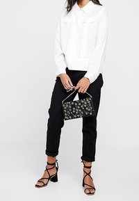 Blouse blanche à manches longues avec col, pantalon noir retroussé et sandales noires à brides. Tenant un sac à main noir à motif floral.