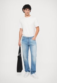 Jeans in denim azzurro con taglio dritto, abbinati a una maglietta bianca semplice. La modella tiene una borsa tote nera e indossa sneakers bianche.