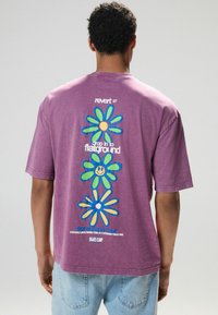 Lila Baumwoll-T-Shirt mit einem bunten Blumenmotiv auf der Rückseite, versehen mit Textelementen, in lockerer Passform und kurzen Ärmeln.