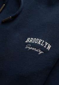 Granatowa bluza wykonana z miękkiego materiału, z białym haftowanym napisem "BROOKLYN" i "Superdry" na klatce piersiowej. Zawiera kaptur ze sznurkiem.