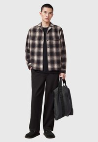 Chemise en flanelle à carreaux marron et noirs, t-shirt noir, pantalon noir lâche, chaussures noires, tenant un sac fourre-tout gris avec un logo.