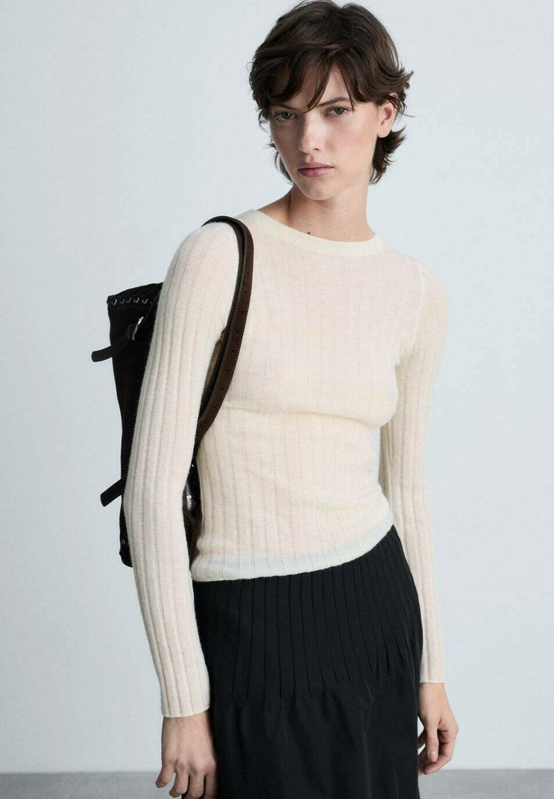 Hellbeige gerippter Pullover mit langen Ärmeln und rundem Halsausschnitt. Getragen mit einem schwarzen Faltenrock und einem dunklen Rucksack.