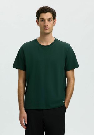 Jeune homme aux cheveux courts et bouclés, debout face à l'objectif, portant un T-shirt uni vert foncé et un pantalon noir sur un fond gris clair.