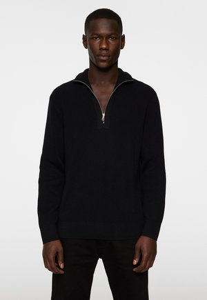 Pull en tricot noir côtelé avec un col zippé quart, doté d'un tissu texturé et d'une coupe décontractée, associé à un pantalon noir.