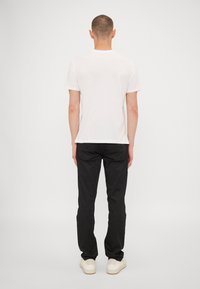 Witte katoenen T-shirt met ronde hals, gecombineerd met zwarte broek en witte sneakers, gezien vanuit de achterkant tegen een effen achtergrond.