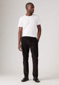 Camiseta blanca de cuello redondo, pantalones de pana negros y zapatos negros. El conjunto es sencillo y bien ajustado, con una paleta de colores neutra.