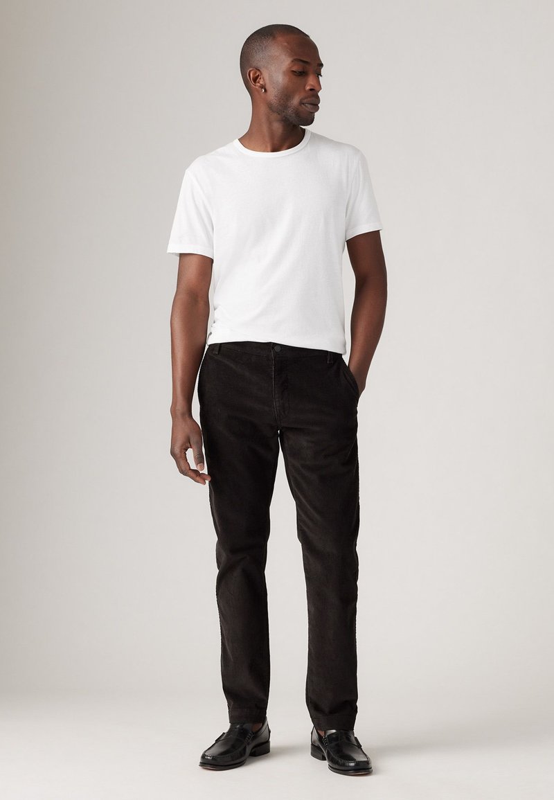 Camiseta blanca de cuello redondo, pantalones de pana negros y zapatos negros. El conjunto es sencillo y bien ajustado, con una paleta de colores neutra.
