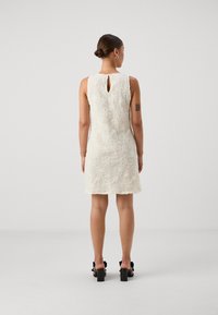 Robe en dentelle blanche avec un motif floral texturé, design sans manches et détails en ouverture dans le dos. Des sandales à talons carrés noires complètent le look.