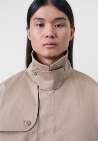 Trench-coat en coton beige avec un col haut, fermeture à boutons-pression et une texture lisse, porté par une personne ayant les cheveux longs et raides.