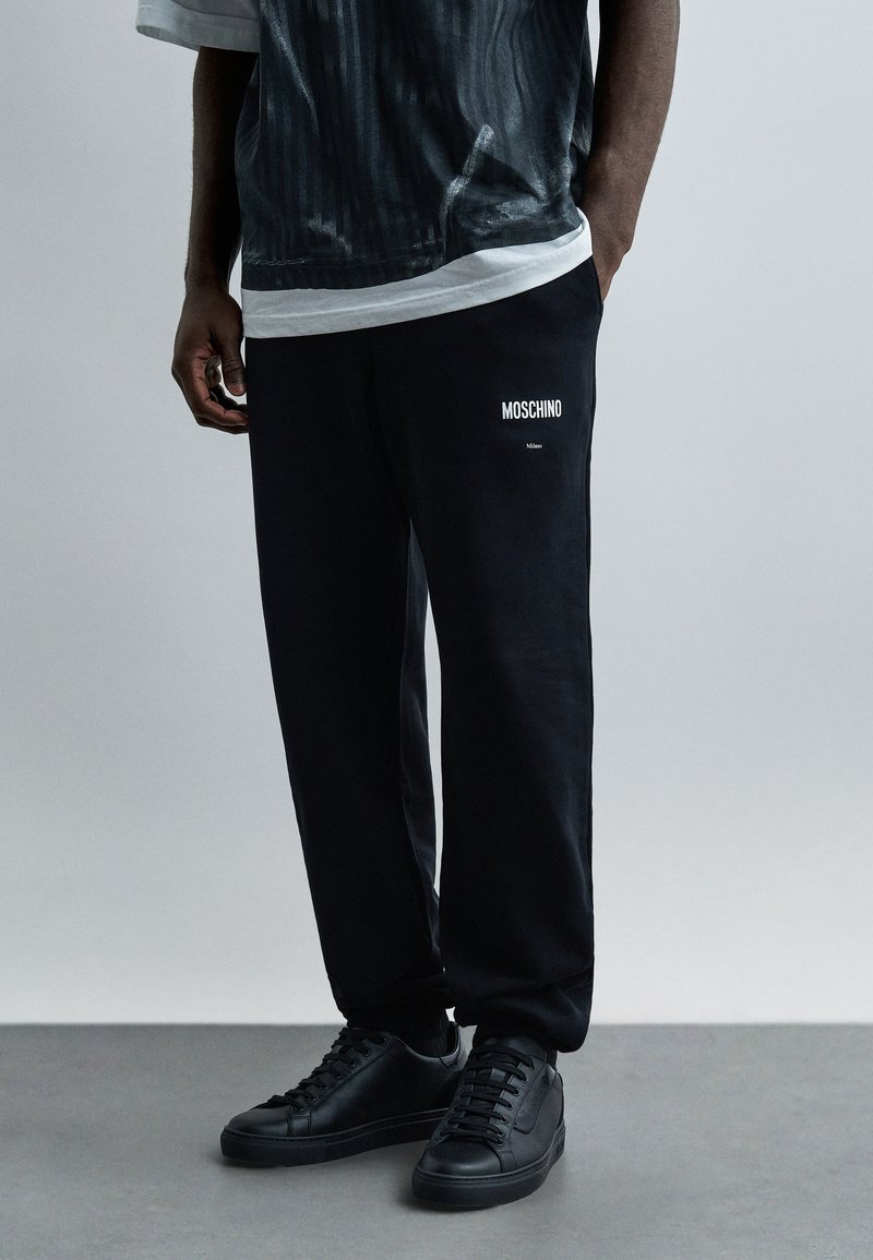 Άνδρας που φοράει μαύρη φόρμα Moschino, μαύρα αθλητικά παπούτσια και σκουρόχρωμο πουκάμισο με σχέδια πάνω από ένα λευκό μπλουζάκι, στέκεται με το ένα χέρι στην τσέπη.