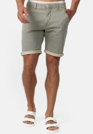 Shorts à carreaux gris clair pour homme avec un ourlet retroussé, dotés d'une fermeture à bouton et à fermeture éclair, fabriqués en tissu respirant. Portés avec des sandales blanches.