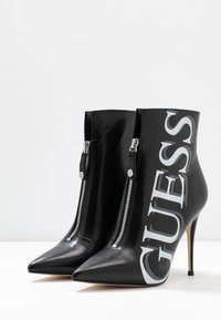 Botas de tobillo negras de tacón alto con punta afilada, textura brillante, cremalleras plateadas y un destacado logo "GUESS" en blanco en el lateral.