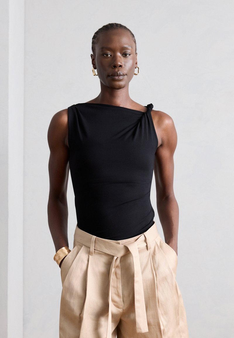 Γυναίκα με κοντά πλεκτά μαλλιά φοράει μαύρο αμάνικο τοπ και μπεζ ψηλόμεση πλισέ παντελόνα με τα χέρια στις τσέπες, μπροστά από απλό φόντο.