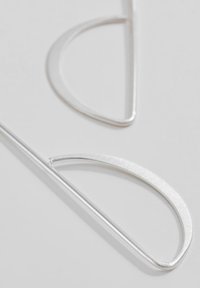 Pendientes de metal plateado con un diseño minimalista, que presentan formas curvadas y una textura suave, posicionados sobre un fondo claro.