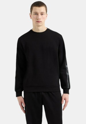 Felpa nera in morbido tessuto con polsini e orlo a coste, presenta un elemento di design grigio a contrasto sulle maniche. Il modello indossa pantaloni neri abbinati.