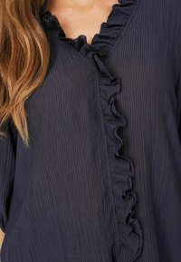 Blusa blu navy con tessuto texturizzato, scollatura a V e un arricciatura decorativa lungo l'apertura frontale. Materiale morbido e leggero.